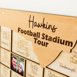 May include: A wooden wall art piece with the words "Hawkins Football Stadium Tour" engraved in a stylish font. The artwork features a map-like design with cutouts for photos and stadium names, including "Gillette Stadium" and "Paycor Stadium."