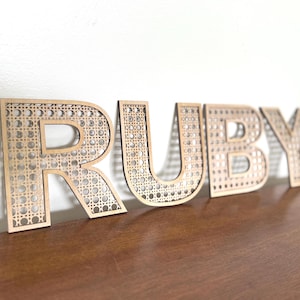 K&ouml;nnte beinhalten: Holzbuchstaben mit dem Schriftzug "RUBY" mit einem geflochtenen Rohrdesign. Die Buchstaben sind hellbraun und stehen auf einer dunkelbraunen Oberfl&auml;che vor einer wei&szlig;en Wand. Die Buchstaben sind mit einem dekorativen Muster ausgeschnitten.