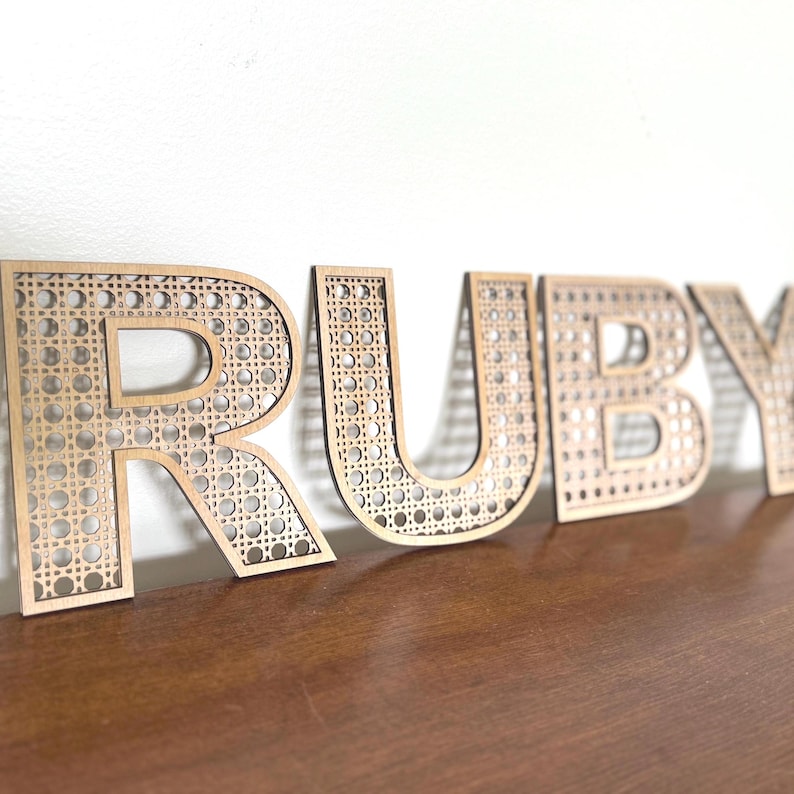 K&ouml;nnte beinhalten: Holzbuchstaben mit dem Schriftzug "RUBY" mit einem geflochtenen Rohrdesign. Die Buchstaben sind hellbraun und stehen auf einer dunkelbraunen Oberfl&auml;che vor einer wei&szlig;en Wand. Die Buchstaben sind mit einem dekorativen Muster ausgeschnitten.
