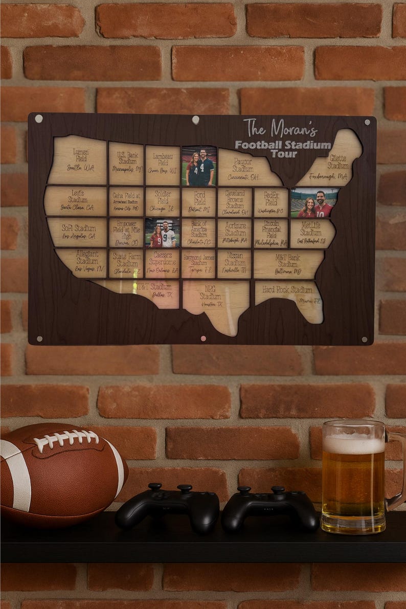 May include: A wooden map of the United States with cutouts for photos and stadium names, labeled "The Moran's Football Stadium Tour." A football, two game controllers, and a beer mug sit below the map. The map is mounted on a brick wall.