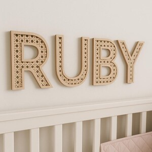 K&ouml;nnte beinhalten: Holzbuchstaben mit der Aufschrift "RUBY" an einer wei&szlig;en Wand. Die Buchstaben sind hellbraun mit einem gewebten Rohrdesign in der Mitte. Die Buchstaben befinden sich &uuml;ber einem wei&szlig;en Kinderbett.