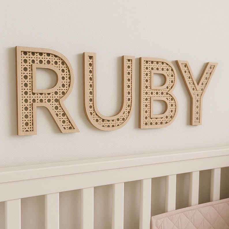 K&ouml;nnte beinhalten: Holzbuchstaben mit der Aufschrift "RUBY" an einer wei&szlig;en Wand. Die Buchstaben sind hellbraun mit einem gewebten Rohrdesign in der Mitte. Die Buchstaben befinden sich &uuml;ber einem wei&szlig;en Kinderbett.