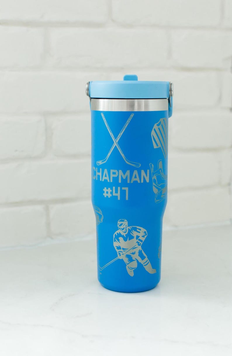May include: A blue insulated tumbler with a light blue lid and silver accents. The tumbler features hockey-themed graphics and the name "Chapman" with the number "47". The tumbler is designed to keep beverages cold or hot.