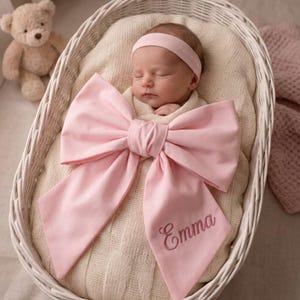 May include: A swaddled infant wearing a light pink headband and a large, matching light pink fabric bow over the swaddle. The bow features the name Emma embroidered in a cursive script on one of the fabric tails, resting in a white basket.