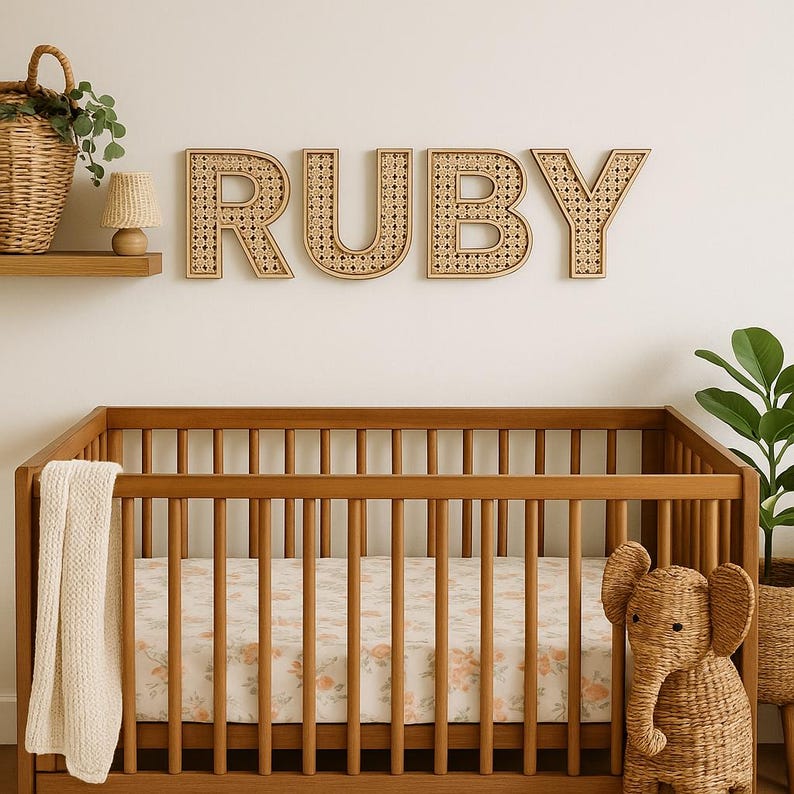 K&ouml;nnte beinhalten: Eine Szene im Kinderzimmer mit einem Holzbett und einem floral gemusterten Laken. &Uuml;ber dem Bett steht der Name "RUBY" in gro&szlig;en Buchstaben aus geflochtenem Rattan. Ein Elefanten-Spielzeug aus Korbgeflecht steht neben dem Bett. Ein geflochtener Korb und eine kleine Lampe stehen auf einem Regal.