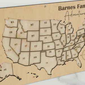 Może przedstawiać: Drewniana mapa Stan&oacute;w Zjednoczonych z konturami i skr&oacute;tami stan&oacute;w. Mapa jest wygrawerowana tekstem "Barnes Family Adventures" i r&oacute;żą wiatr&oacute;w. Mapa jest zamontowana na jasnej drewnianej desce.