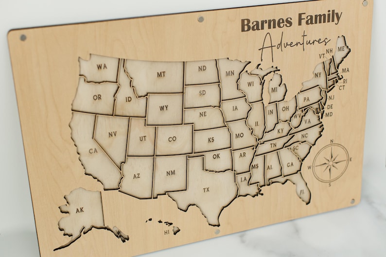 Może przedstawiać: Drewniana mapa Stan&oacute;w Zjednoczonych z konturami i skr&oacute;tami stan&oacute;w. Mapa jest wygrawerowana tekstem "Barnes Family Adventures" i r&oacute;żą wiatr&oacute;w. Mapa jest zamontowana na jasnej drewnianej desce.