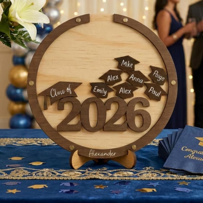 May include: A wooden graduation sign with a circular frame and a light wood center. The sign features the text "Class of 2026" and individual graduation caps with names. A banner at the bottom reads "Alexander".