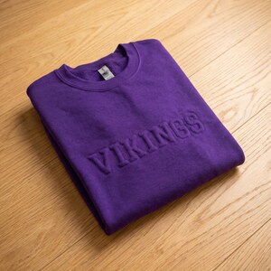 Puede incluir: Una sudadera morada doblada con la palabra "VIKINGS" en relieve en la parte delantera. La sudadera est&aacute; hecha de un material suave y se muestra sobre una superficie de madera de color claro.