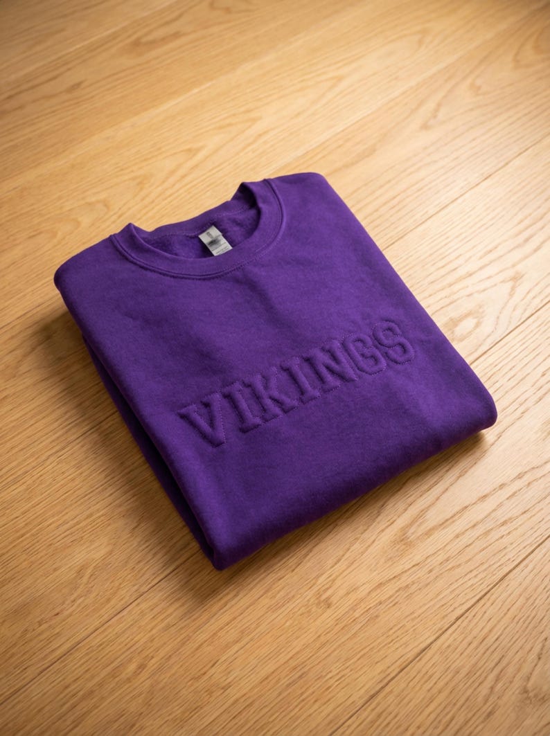 Puede incluir: Una sudadera morada doblada con la palabra "VIKINGS" en relieve en la parte delantera. La sudadera est&aacute; hecha de un material suave y se muestra sobre una superficie de madera de color claro.