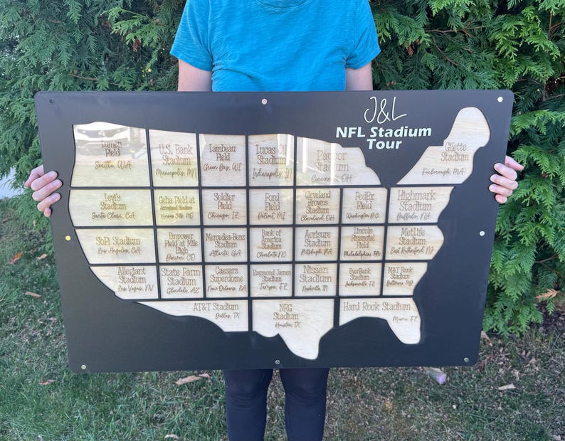 May include: A black wooden sign with a map of the United States, featuring cutouts for NFL stadiums. The sign includes the text "J&L NFL Stadium Tour" and stadium names. The sign is held up by a person.