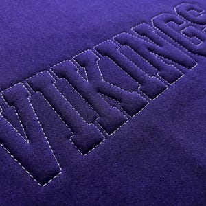 Puede incluir: Primer plano de una tela morada oscura con la palabra "VIKINGS" cosida en letras de bloque en relieve. Las letras est&aacute;n delineadas con costuras blancas, creando un efecto de contraste. La tela tiene un aspecto suave y texturizado.