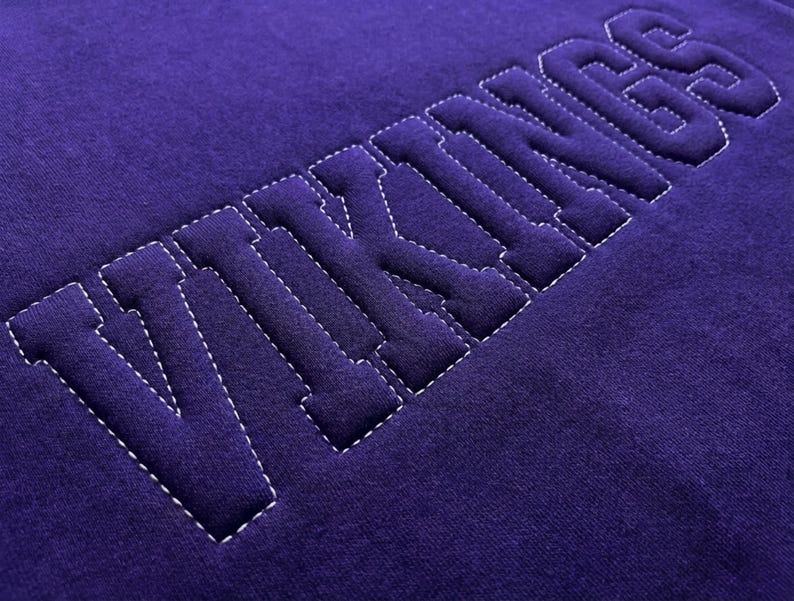 Puede incluir: Primer plano de una tela morada oscura con la palabra "VIKINGS" cosida en letras de bloque en relieve. Las letras est&aacute;n delineadas con costuras blancas, creando un efecto de contraste. La tela tiene un aspecto suave y texturizado.
