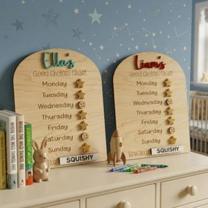 Puede incluir: Dos tablas de buenas elecciones de madera con nombres en letras coloridas. Cada tabla tiene una lista de d&iacute;as con pegatinas de estrellas y caras sonrientes. Un cohete de madera, crayones y una figura de conejito est&aacute;n sobre la mesa.