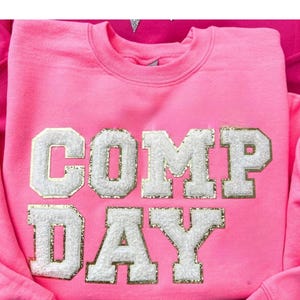 May include: Pink sweatshirt with the words "COMP DAY" in white letters with gold glitter accents.
