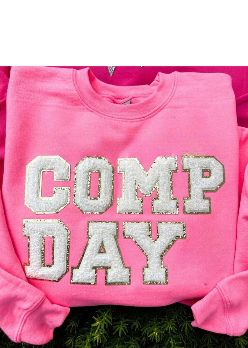 May include: Pink sweatshirt with the words "COMP DAY" in white letters with gold glitter accents.