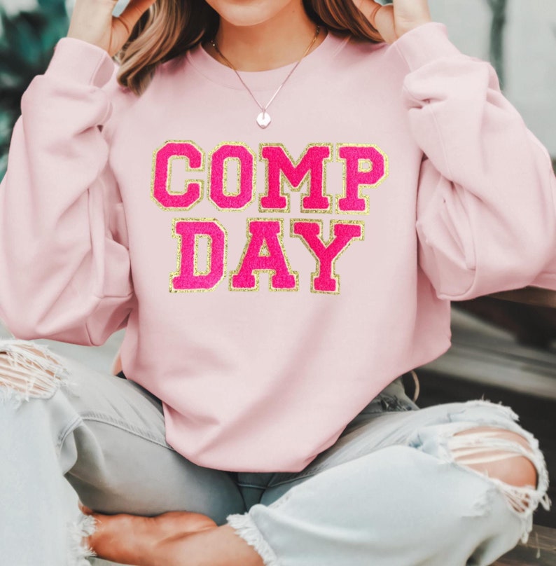 May include: A pink sweatshirt with the text "COMP DAY" in gold glitter lettering.