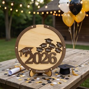 May include: A wooden graduation sign with the text "Class of 2026" and names of graduates. The sign is round with a dark brown frame and a light wood center. A graduation cap and confetti are on the table.
