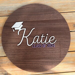 May include: A round wooden sign with a white graduation cap and the name "Katie" in white script. The text "Class of 2024" is written in purple below the name.