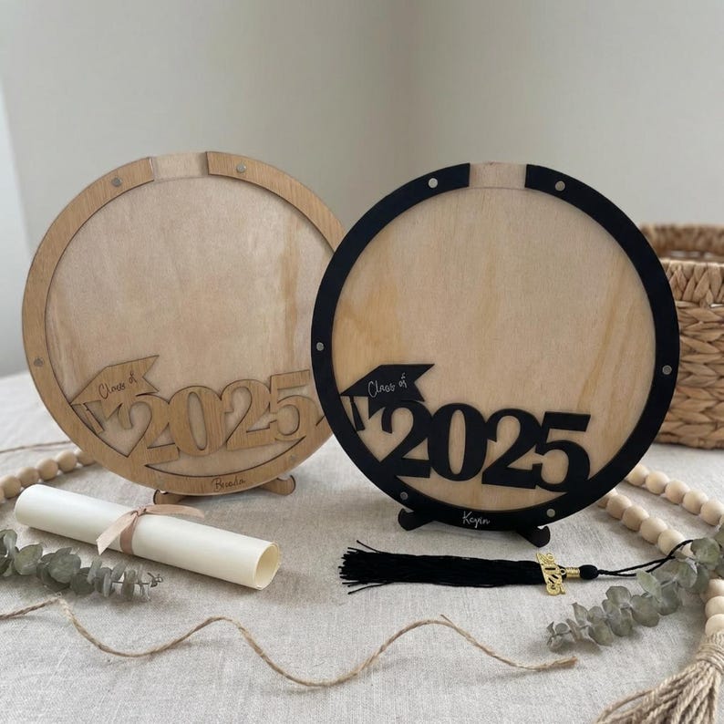 May include: Two wooden graduation frames with the text "Class of 2025". One frame is natural wood, the other is black. A diploma and tassel are also present. The frames are round with a stand.