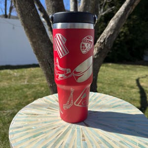 May include: A red insulated tumbler with a black lid and silver handle. The tumbler features etched hockey-themed graphics, including a jersey, helmet, skates, puck, and hockey sticks. The tumbler is cylindrical and sits on a mosaic table.