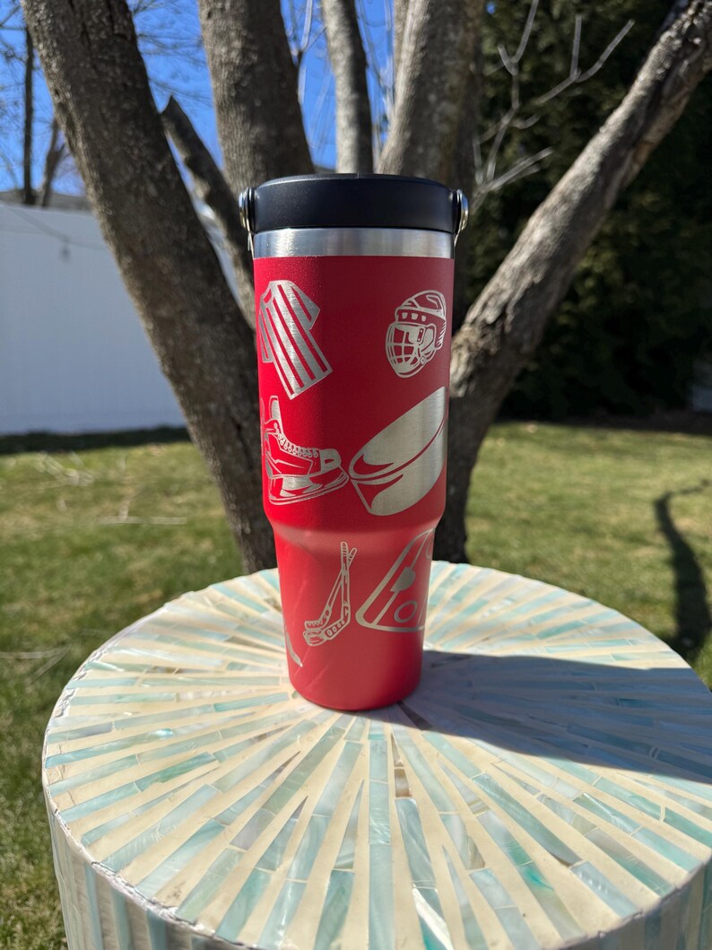 May include: A red insulated tumbler with a black lid and silver handle. The tumbler features etched hockey-themed graphics, including a jersey, helmet, skates, puck, and hockey sticks. The tumbler is cylindrical and sits on a mosaic table.