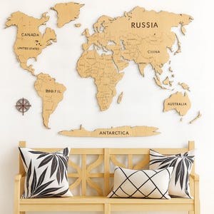 May include: A wooden world map with country names is mounted on a white wall. Below the map is a light wood bench with three decorative cushions. The cushions have black and white geometric and leaf patterns. A compass is mounted on the wall.