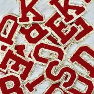 May include: A collection of red fabric letters with gold trim. The letters are in a varsity style, with a thick, textured appearance. The letters are scattered on a white surface.