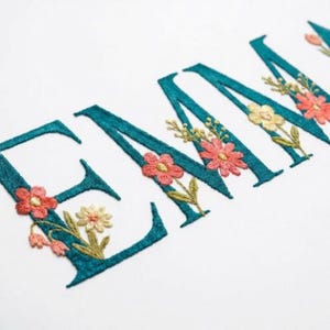 May include: Teal embroidered letters spelling "EMMA" with floral accents. The flowers are in shades of coral, yellow, and green. The embroidery is on a white background, creating a personalized and decorative design.
