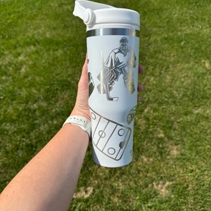 May include: A white insulated tumbler with a silver rim and a white lid. The tumbler features silver graphics of a hockey goalie, hockey stick, and a hockey rink. The tumbler is held in a person's hand against a green grass background.