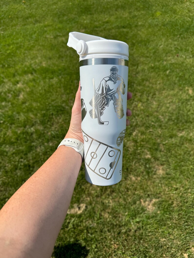 May include: A white insulated tumbler with a silver rim and a white lid. The tumbler features silver graphics of a hockey goalie, hockey stick, and a hockey rink. The tumbler is held in a person's hand against a green grass background.