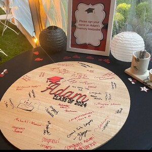 May include: A round wooden sign with the words "Adam Class of 2024" in red lettering. The sign is covered in signatures and well wishes for the graduate. The sign is on a black table with a white paper lantern and a small ceramic cup and saucer.
