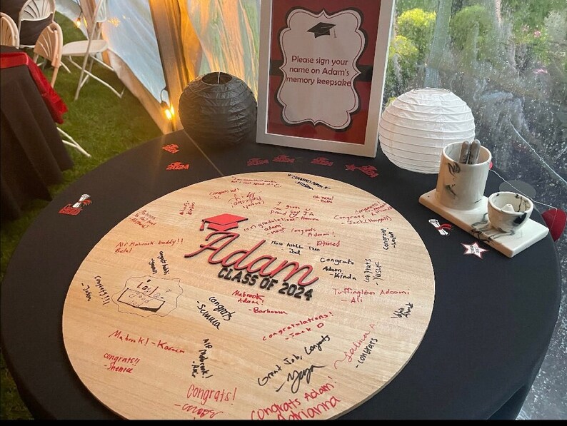May include: A round wooden sign with the words "Adam Class of 2024" in red lettering. The sign is covered in signatures and well wishes for the graduate. The sign is on a black table with a white paper lantern and a small ceramic cup and saucer.