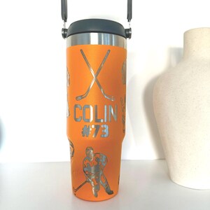 May include: An orange insulated tumbler with a black handle and lid. The tumbler features silver hockey-themed graphics, including crossed hockey sticks, a hockey player, and the name "COLIN #73". The tumbler is designed for cold and hot beverages.