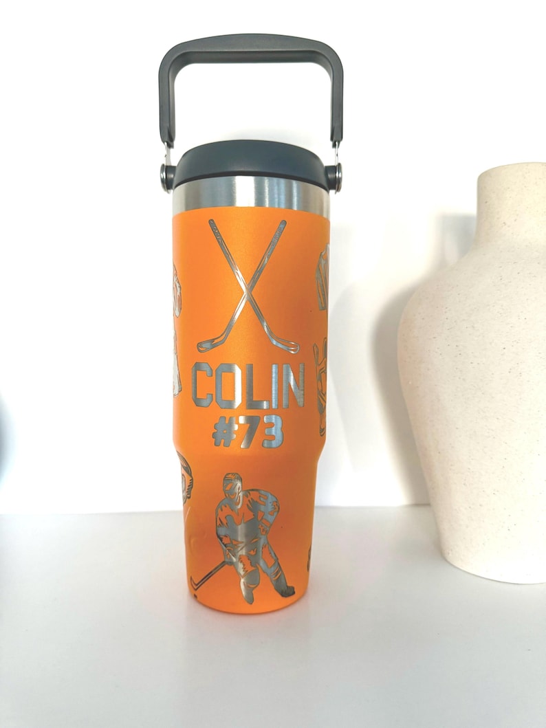 May include: An orange insulated tumbler with a black handle and lid. The tumbler features silver hockey-themed graphics, including crossed hockey sticks, a hockey player, and the name "COLIN #73". The tumbler is designed for cold and hot beverages.