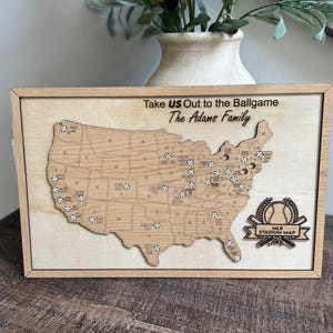 May include: Wooden MLB stadium map with a light wood frame. The map features a laser-cut design of the United States with baseball icons marking stadium locations. Text includes "Take US Out to the Ballgame" and "The Adams Family". The bottom reads "MLB STADIUM MAP".