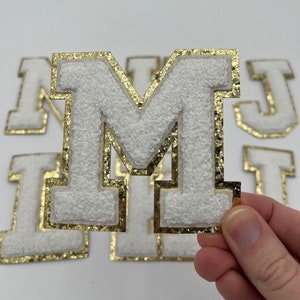 May include: White felt letter patches with gold glitter trim. The letters are M, I, and J.