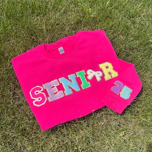 May include: Pink sweatshirt with the word "SENIOR" in rainbow glitter letters and the number "23" on the sleeve.