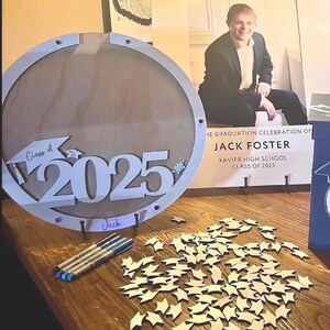 May include: A wooden graduation sign with the text "Class of 2025" and the name "Jack". The sign is round with a light wood finish. There are several small wooden star cutouts and pens on the table. A poster with the word "welcome" is in the background.