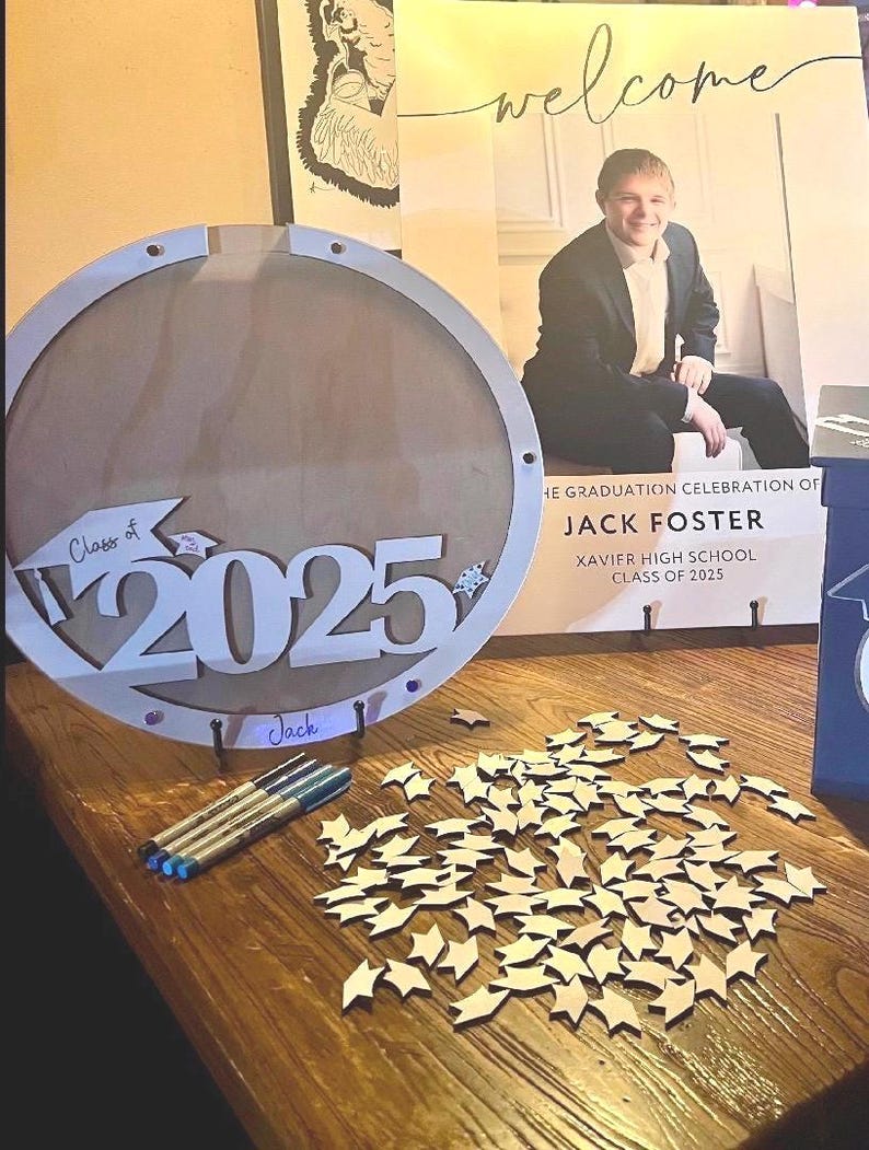 May include: A wooden graduation sign with the text "Class of 2025" and the name "Jack". The sign is round with a light wood finish. There are several small wooden star cutouts and pens on the table. A poster with the word "welcome" is in the background.