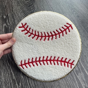 May include: White and red baseball-themed round rug with a gold trim. The rug is made of a fluffy, textured material and features red stitching.