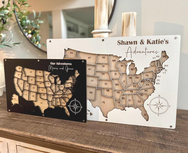 Może przedstawiać: Dwie drewniane tablice z mapą Stan&oacute;w Zjednoczonych. Jedna jest czarna z napisem "Our Adventures Morris and Grace" i kompasem. Druga jest biała z napisem "Shawn & Katie's Adventures" i kompasem.