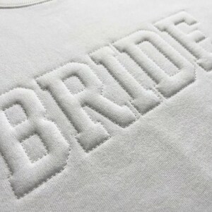 Puede incluir: Primer plano de una sudadera blanca con la palabra "BRIDE" bordada en relieve, con una fuente texturizada. Las letras son en negrita, de estilo bloque, creando un efecto 3D. La tela tiene un aspecto suave y ligeramente texturizado.