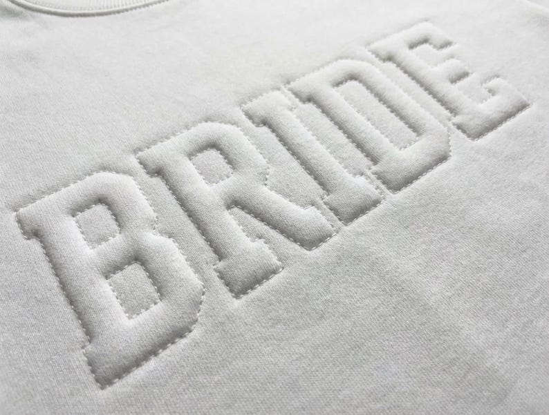 Puede incluir: Primer plano de una sudadera blanca con la palabra "BRIDE" bordada en relieve, con una fuente texturizada. Las letras son en negrita, de estilo bloque, creando un efecto 3D. La tela tiene un aspecto suave y ligeramente texturizado.