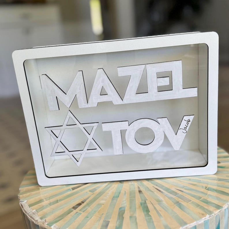 Peut inclure: Une bo&icirc;te en bois blanche avec les mots "MAZEL TOV" d&eacute;coup&eacute;s dans une police sans-serif en gras. Une &eacute;toile de David est repr&eacute;sent&eacute;e sous le mot "MAZEL". Le nom "Jacob" est &eacute;crit en cursive sur le c&ocirc;t&eacute; droit. La bo&icirc;te est pr&eacute;sent&eacute;e sur une surface d&eacute;corative.