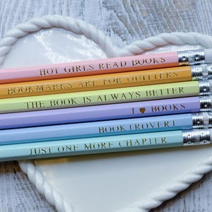May include: A collection of pastel-colored pencils with text, including "Hot Girls Read Books" and "Just One More Chapter." The pencils are arranged on a white heart-shaped dish with a rope-like border. The pencils have silver metal ends.