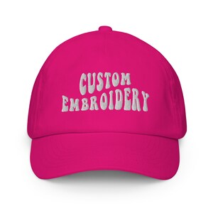 Peut inclure: Une casquette de baseball rose avec un texte brodé blanc qui dit "CUSTOM EMBROIDERY".