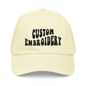 Può includere: Un berretto da baseball giallo chiaro con testo ricamato in nero che recita "CUSTOM EMBROIDERY".