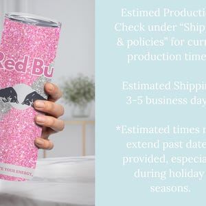 May include: A pink glitter tumbler featuring the Red Bull logo in white and black. The tumbler has the text "Red Bu" and "IGNITE YOUR ENERGY" printed on it. The tumbler is held by a person's hand.