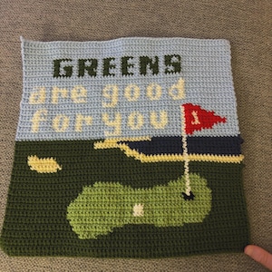 May include: Crocheted golf-themed wall hanging. The top section is light blue with the words "GREENS are good for you" in green and yellow. The bottom section depicts a green golf course with a red flag. A unique piece for golf enthusiasts.